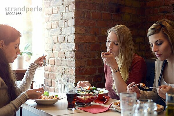 Drei attraktive junge Freundinnen entspannen sich beim gemeinsamen Mittagessen in einem Restaurant  sitzen an einem Tisch  unterhalten sich und genießen ihr Essen