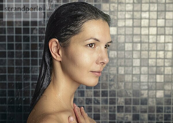 Attraktive Frau  die sich unter der Dusche die Haare wäscht und sie unter dem Wasserstrahl abspült und dabei entspannt wegschaut  in einem Konzept für Haarpflege  Schönheit und Hygiene