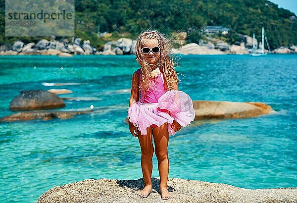 Nettes kleines Mädchen in Sonnenbrille und rosa Kleid posiert auf schönen tropischen Strand. Sommer  Spaß und Urlaub Konzept