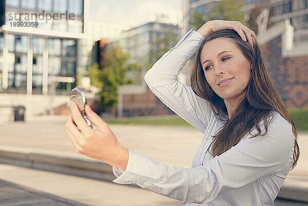 Schöne junge Frau  die ihr Aussehen in einem kleinen Handspiegel auf der Straße überprüft und lächelt  während sie ihre langen braunen Haare aus dem Gesicht hält