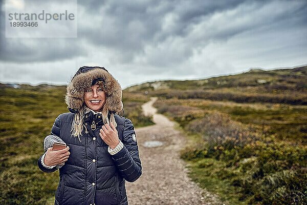 Frau mittleren Alters trotzt einem kalten Wintertag in der Natur und spaziert an einem windigen Tag durch die Landschaft und lächelt glücklich in die Kamera