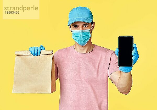 Ein Zusteller hält ein Papierpaket mit Lebensmitteln und einem Smartphone in der Hand und trägt einen Mundschutz und Handschuhe zum Schutz vor dem Coronavirus COVID 19. Zustelldienst mit moderner Technologie