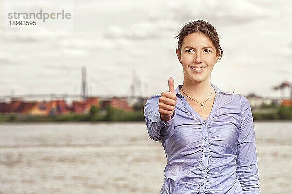 Glücklich lächelnde attraktive Frau  die einen Daumen nach oben Geste des Erfolgs und der Zustimmung  Oberkörper Porträt vor einem städtischen Fluss oder Wasserstraße mit copyspace