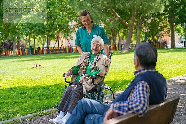Eine ältere Frau mit der Krankenschwester auf einem Spaziergang im Garten eines Pflegeheims in einem Rollstuhl in der Natur und begrüßt einen älteren Mann
