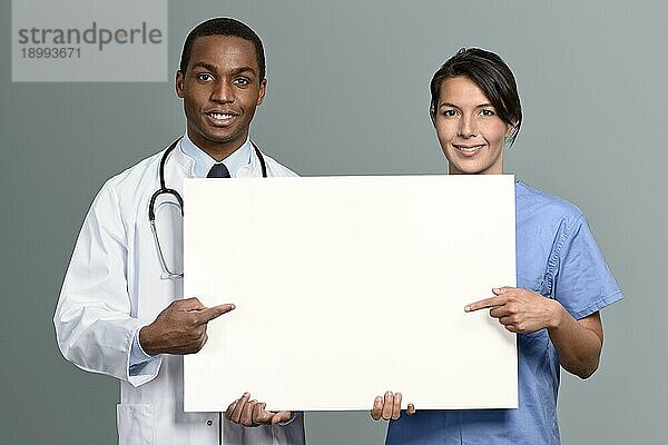 Multiethnisches Ärzteteam mit einem afrikanischen Arzt im Laborkittel und Stethoskop und einer Krankenschwester in OP Kleidung  die ein leeres weißes Schild halten  auf dem Ihr Text mit einem freundlichen Lächeln zu lesen ist