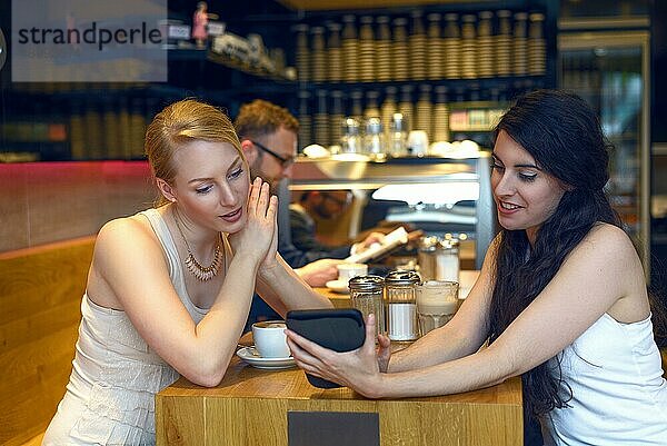 Zwei junge Frauen schauen auf ein digitales Tablet  während sie in einem Bistro oder einer Kaffeebar zusammensitzen und einen Kaffee genießen