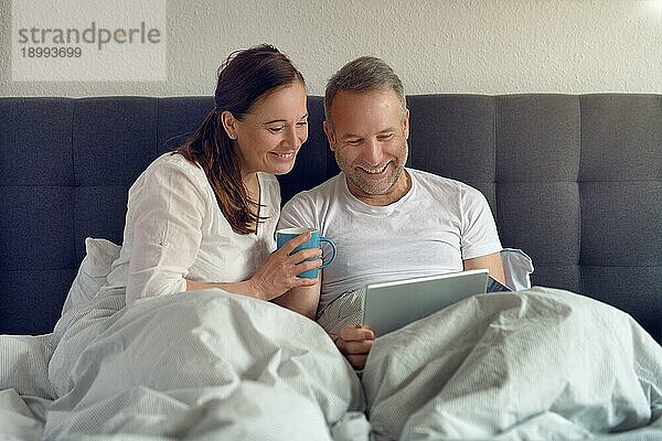 Glückliches Paar mittleren Alters  das morgens im Bett sitzt  Kaffee trinkt und mit einem digitalen Tablet online einkauft