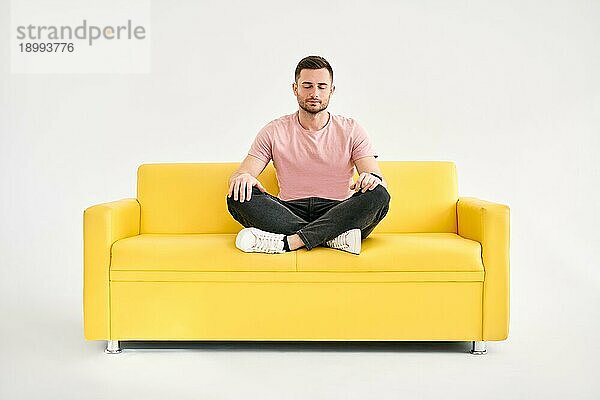 Hübscher junger Mann sitzt in Yogalotuspose und meditiert auf einem bequemen gelben Sofa vor weißem Hintergrund