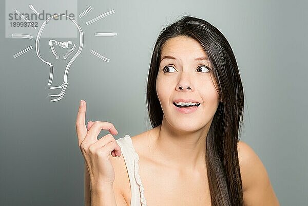 Attraktive junge Frau signalisiert mit ihrem Zeigefinger eine Idee in Form einer illustrierten Glühbirne  auf einem grauen Studiohintergrund