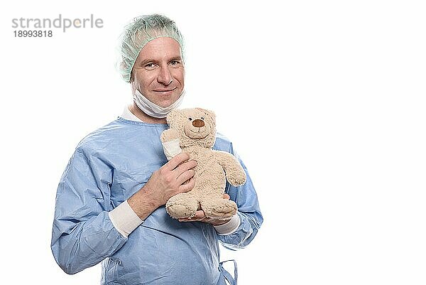 Freundlich lächelnder Kinderarzt mittleren Alters in blauem Kittel  der einen Teddybär mit bandagiertem Arm hält  um einen jungen Patienten zu beruhigen  vor weißem Hintergrund