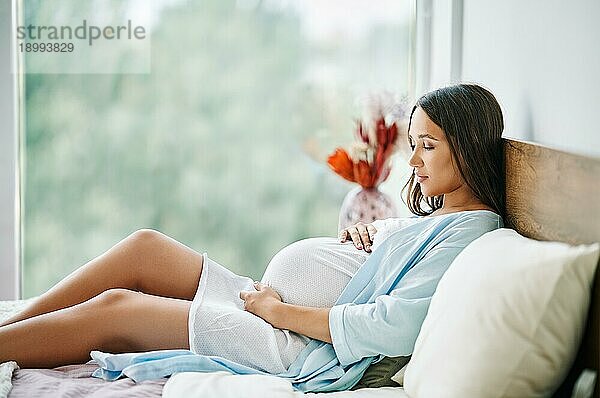 Portrait einer glücklichen schwangeren Frau  die im Bett liegt und ihren Bauch zu Hause berührt. Mutterschaft  Menschen Konzept. Tender Stimmung Foto der Schwangerschaft