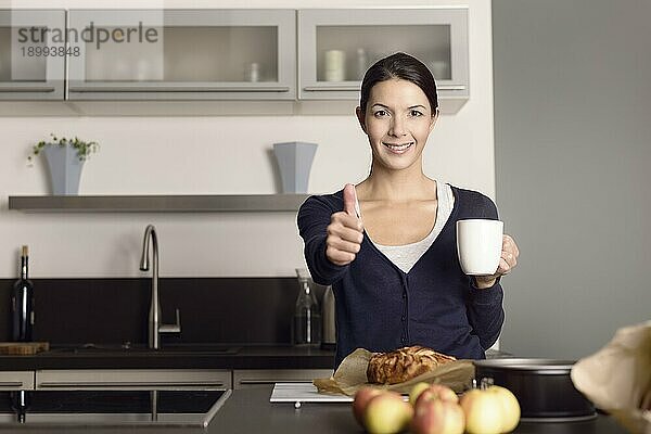 Glückliche attraktive junge Köchin  die den Daumen nach oben streckt  während sie in ihrer Küche steht und Kaffee trinkt  während sie einen frisch gebackenen Apfelkuchen vor sich hat