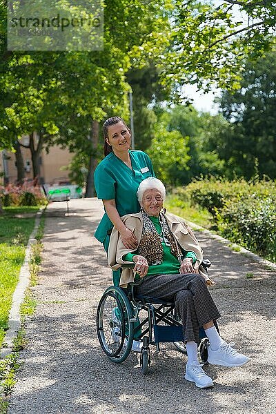 Porträt einer älteren Frau mit der Krankenschwester bei einem Spaziergang im Garten eines Pflegeheims im Rollstuhl