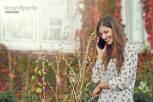 Lächelnde junge Frau  die mit einem Handy im Herbst vor einem weißen Haus mit bunten Kletterpflanzen steht und lächelt