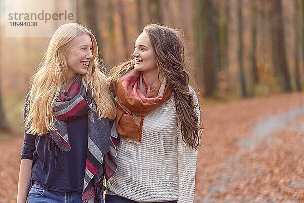 Zwei attraktive junge Freundinnen spazieren Arm in Arm durch einen herbstlichen Wald  plaudernd und sich gegenseitig anlächelnd  Nahaufnahme des Oberkörpers