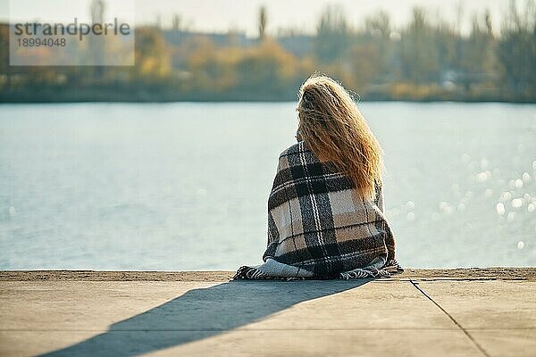 Rückenansicht einer jungen Frau  die sich allein an einem Fluss in der Stadt ausruht  eingewickelt in eine Wolldecke  und die Natur genießt. Ruhe  Reisen Konzept