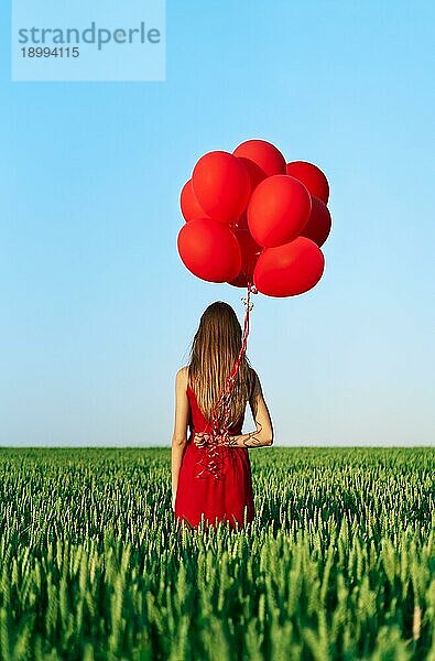 Rückenansicht einer jungen Frau in rotem Kleid  die mit roten Luftballons auf einer grünen Sommerwiese steht. Spaß  Glück  Feier Konzept