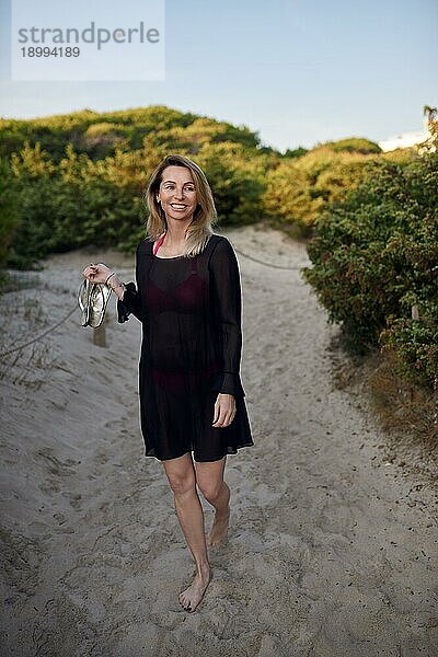 Attraktive schlanke blonde Frau  die ihre Schuhe trägt  während sie auf einem Sandstrandweg spazieren geht und fröhlich zur Seite lächelt  im Sommerurlaub