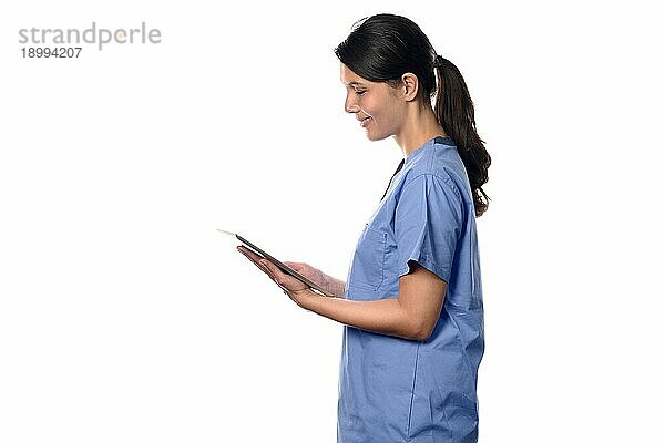 Fröhliche junge brünette Krankenschwester oder Ärztin in blaür Uniform lächelt  während sie ein dünnes drahtloses PC Tablet benutzt  das mit dem Internet verbunden ist  Seitenporträt vor weißem Hintergrund