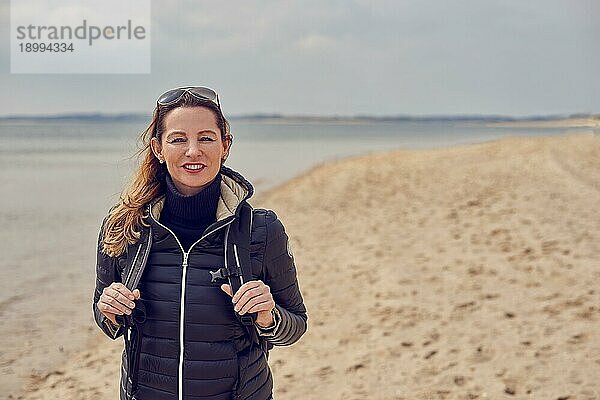 Attraktive  gesunde Frau  die an einem kalten  bewölkten Tag eine Strandwanderung genießt und lächelnd in die Kamera schaut  während sie die Riemen ihres Rucksacks hält