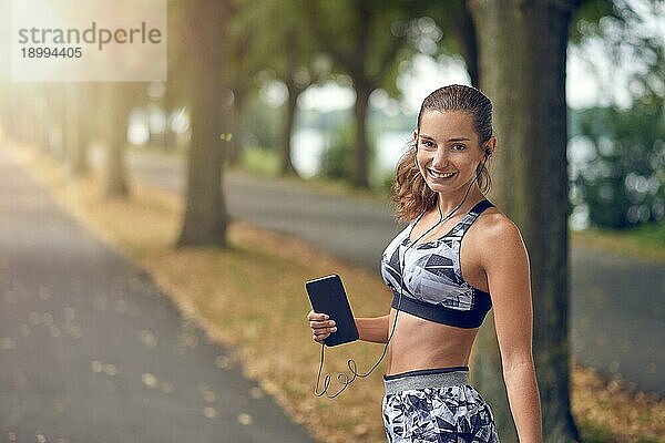 Attraktive sportliche Frau  die mit ihrem Handy Musik hört  joggt auf einer von Bäumen gesäumten Straße und lächelt fröhlich in die Kamera