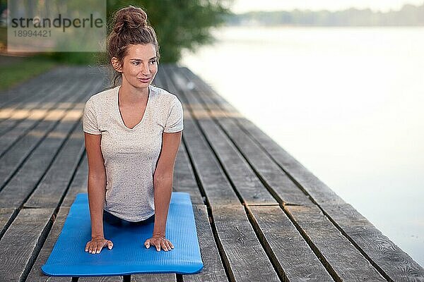 Attraktive junge Frau  die auf einer Matte auf einem Holzdeck über einem See oder Fluss trainiert und dabei eine Yogapose einnimmt und lächelnd auf den See blickt