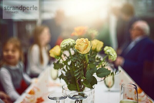 Vase mit frischen gelben und orangefarbenen Blumen als Tischdekoration bei einer Dinnerparty mit selektivem Fokus auf das Arrangement und unscharfen Personen
