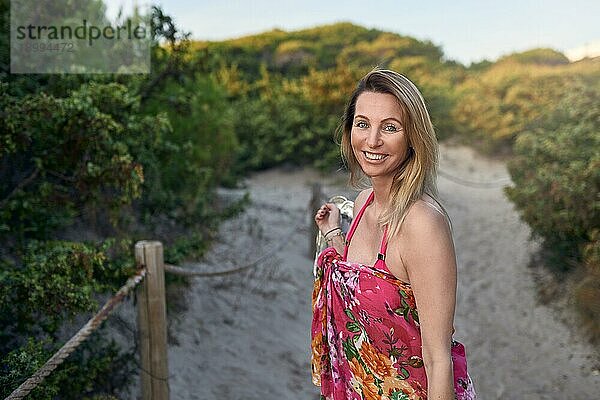 Lachende  fröhliche  temperamentvolle Frau im Sommerurlaub  die ihre Schuhe in der Hand hält  während sie am Strand entlang eines Weges durch den Busch spaziert