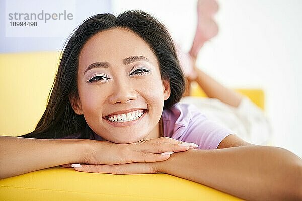 Closeup glücklich asiatische Frau Porträt entspannt zu Hause liegen auf gelben Sofa. Ruhe  weibliche Schönheit