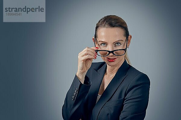 Stilvolle Geschäftsfrau  die mit nachdenklichem  ernstem Gesichtsausdruck über ihre Brille in die Kamera blickt  vor einem grau abgestuften Hintergrund