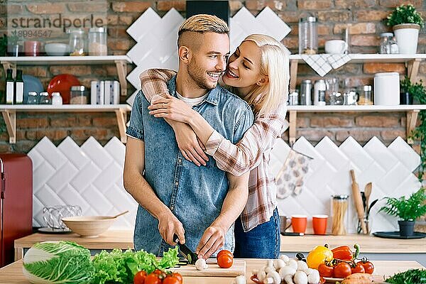 Lächelndes junges Paar kocht zusammen vegetarische Mahlzeit in der Küche zu Hause. Frau umarmt Mann. Gesunder Lebensstil Konzept