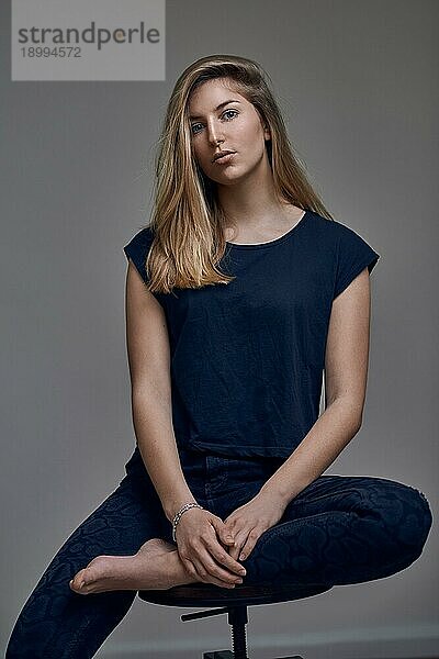 Schöne junge Frau in blauem TShirt mit glattem blondem Haar  die mit einem Bein auf einem Stuhl sitzt und in die Kamera schaut. Front low angle portrait gegen grauen Hintergrund