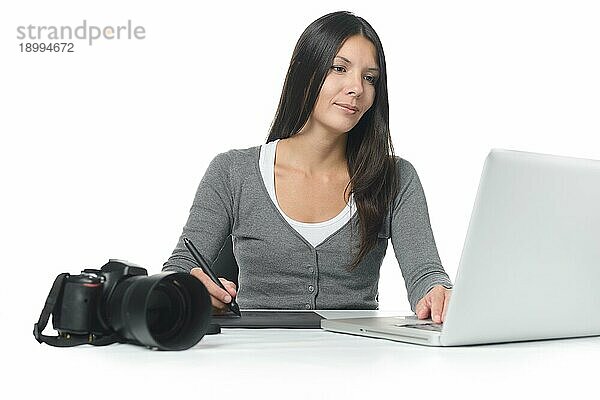 Weibliche Fotografin lächelt  während sie ihre kürzlich heruntergeladenen Dateien auf einem Laptop mit einem Tablet und einem Stift bearbeitet  während ihre Kamera daneben auf dem Schreibtisch liegt  über weiß