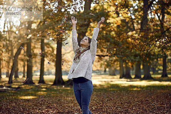 Fröhliche attraktive junge Frau  die mit einem glücklichen Lächeln im Freien in einem Park mit buntem Herbstlaub an den Bäumen Herbstblätter in die Luft wirft