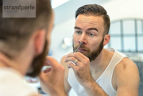 Gutaussehender bärtiger junger Mann  der sich vor dem Spiegel im Badezimmer bei der täglichen Körperpflege die Nasenhaare zupft