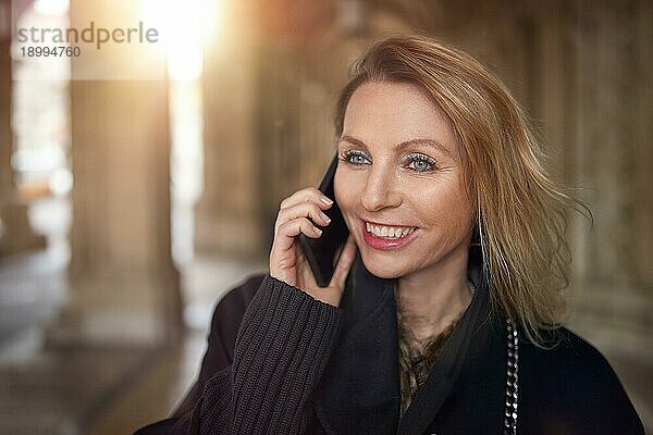 Attraktive Frau spricht mit einem Smartphone in einem großen Gebäude und lächelt  während sie dem Gespräch zuhört