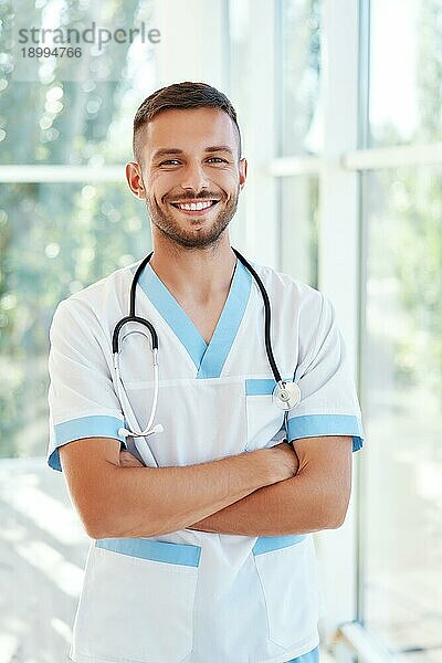 Porträt eines zuversichtlich lächelnden männlichen Arztes mit Stethoskop in medizinischer Uniform  die Arme verschränkt