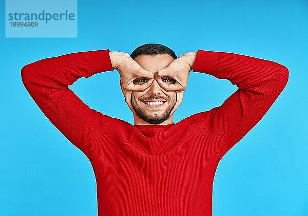 Porträt von glücklichen kreativen Mann macht Finger Gläser auf blauem Hintergrund Studio. Spaß und Menschen Konzept