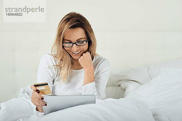 Attraktive Frau entspannt sich im Bett beim Onlineshopping und hält ihre Kreditkarte in der einen Hand und ihr digitales Tablet in der anderen  während sie in die Kamera lächelt