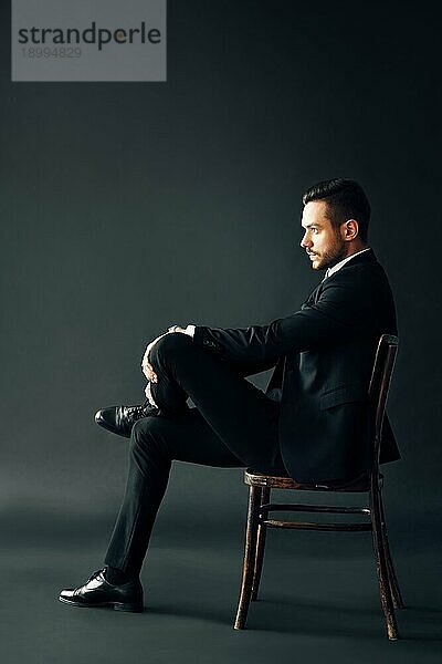 Selbstbewusster  gutaussehender Männchen im schwarzen Anzug  der auf einem Stuhl vor dunklem Hintergrund sitzt. Profilansicht voll lenght männliches Porträt