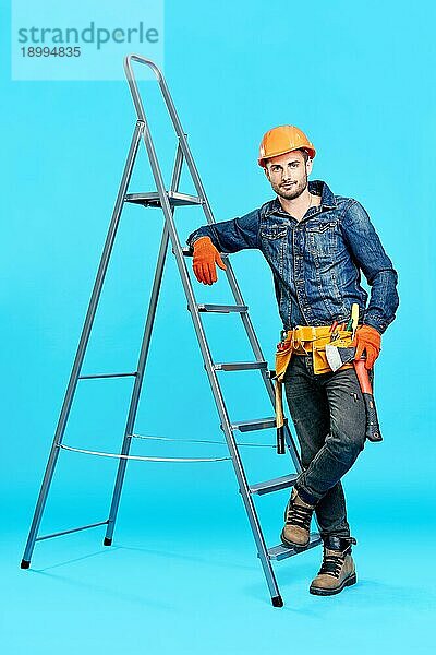 Ganzkörperporträt eines gut aussehenden männlichen Bauarbeiters  der auf einer Leiter über einem blaün Hintergrund klettert und zur Kamera schaut