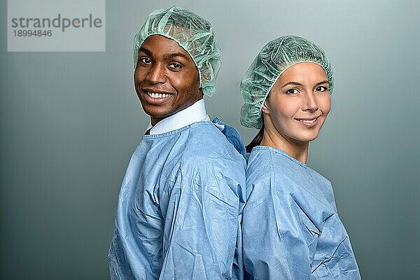 Hübscher junger afrikanischer Arzt oder Krankenpfleger in Kittel und Sterilitätskappe  der direkt in die Kamera schaut  vor grauem Hintergrund  Kopf Schulter Porträt