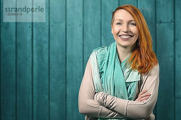 Lächelnde  glückliche  hübsche  rothaarige Frau mit langen  über eine Schulter fallenden Haaren in einem grauen Trikot  die mit verschränkten Armen vor einem Hintergrund mit Kreidetafel und Copyspace steht