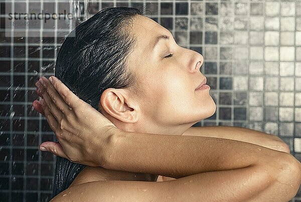 Attraktive Frau wäscht ihr Haar in der Dusche und spült es unter dem Wasserstrahl ab  während sie den Kopf zurücklehnt und die Augen schließt