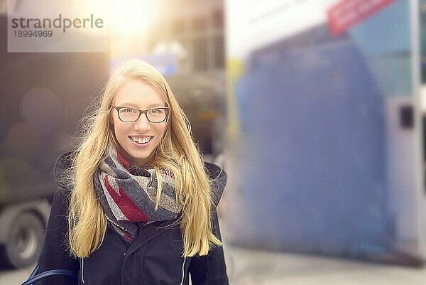 Lächelnde attraktive blonde Frau mit Brille in warmer Wintermode steht in einer städtischen Straße mit heller Sonne Fackel über ihrem Kopf
