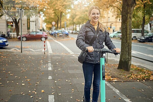 Junge glückliche blonde Frau fährt mit einem Elektroroller in der Stadt  lächelt in die Kamera  im Herbst