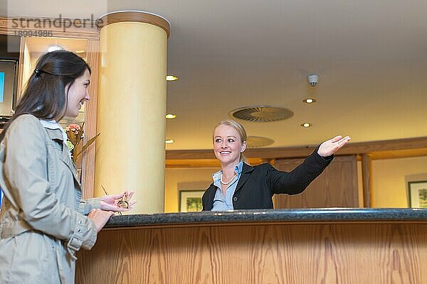 Niedriger Blickwinkel auf eine schöne  freundlich lächelnde Empfangsdame hinter dem Serviceschalter in einer Hotellobby  die einem attraktiven weiblichen Gast mit ihrer Hand den Weg zu ihrer Unterkunft weist
