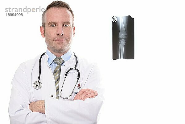 Selbstbewusster männlicher Arzt in Laborkittel und Stethoskop  der mit verschränkten Armen vor einer Röntgenaufnahme eines Kniegelenks steht und in die Kamera schaut  vor weißem Hintergrund