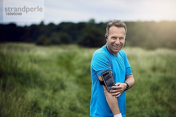 Fröhlicher reifer Mann mit Smartphone am Arm  der auf einer Wiese steht und Musik hört