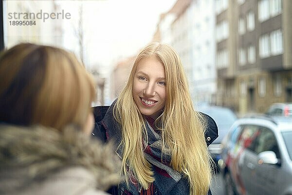 Attraktive junge blonde Frau im Gespräch mit einer Freundin im Freien auf einer belebten städtischen Straße  die lächelt  während sie ihrem Gespräch zuhört